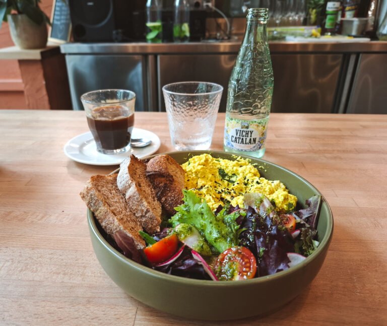 A plate of tofu scramble with fresh salad and bread, served on a wooden counter at Morgentau in Barcelona. In the background, there is a bottle of Vichy Catalan mineral water, a glass of iced coffee, and a glass of water. The setting includes a cozy bar area with an inviting atmosphere.